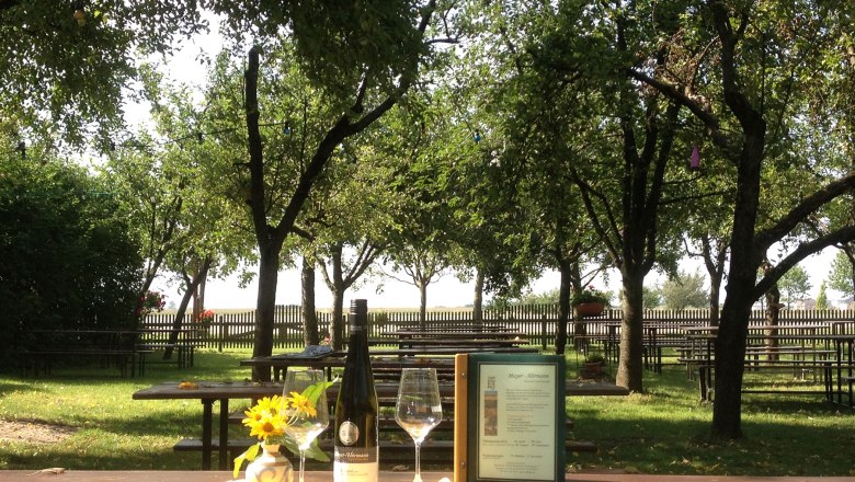 A guest garden with wooden tables, trees and a bottle of wine on a table.