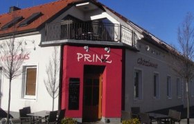 Exterior view of a building with the inscription 'Prinz', guest garden with tables and chairs, blue sky.
