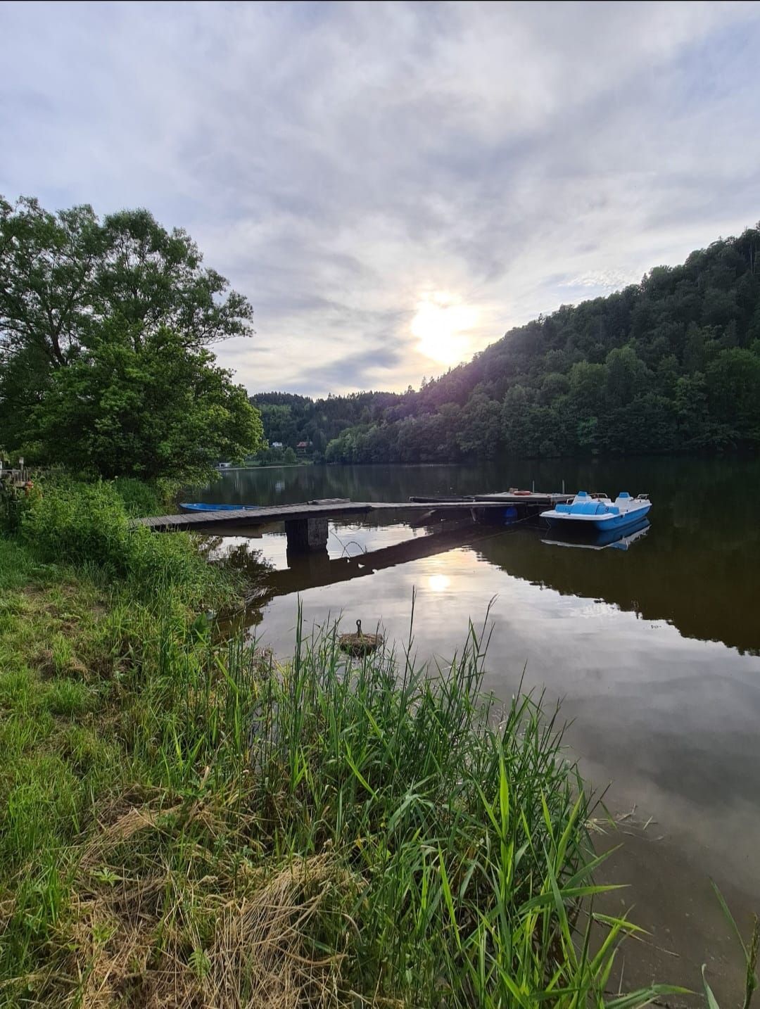 A quiet reservoir with a jetty and a pedal boat in the water, surrounded by green trees and a cloudy sky.