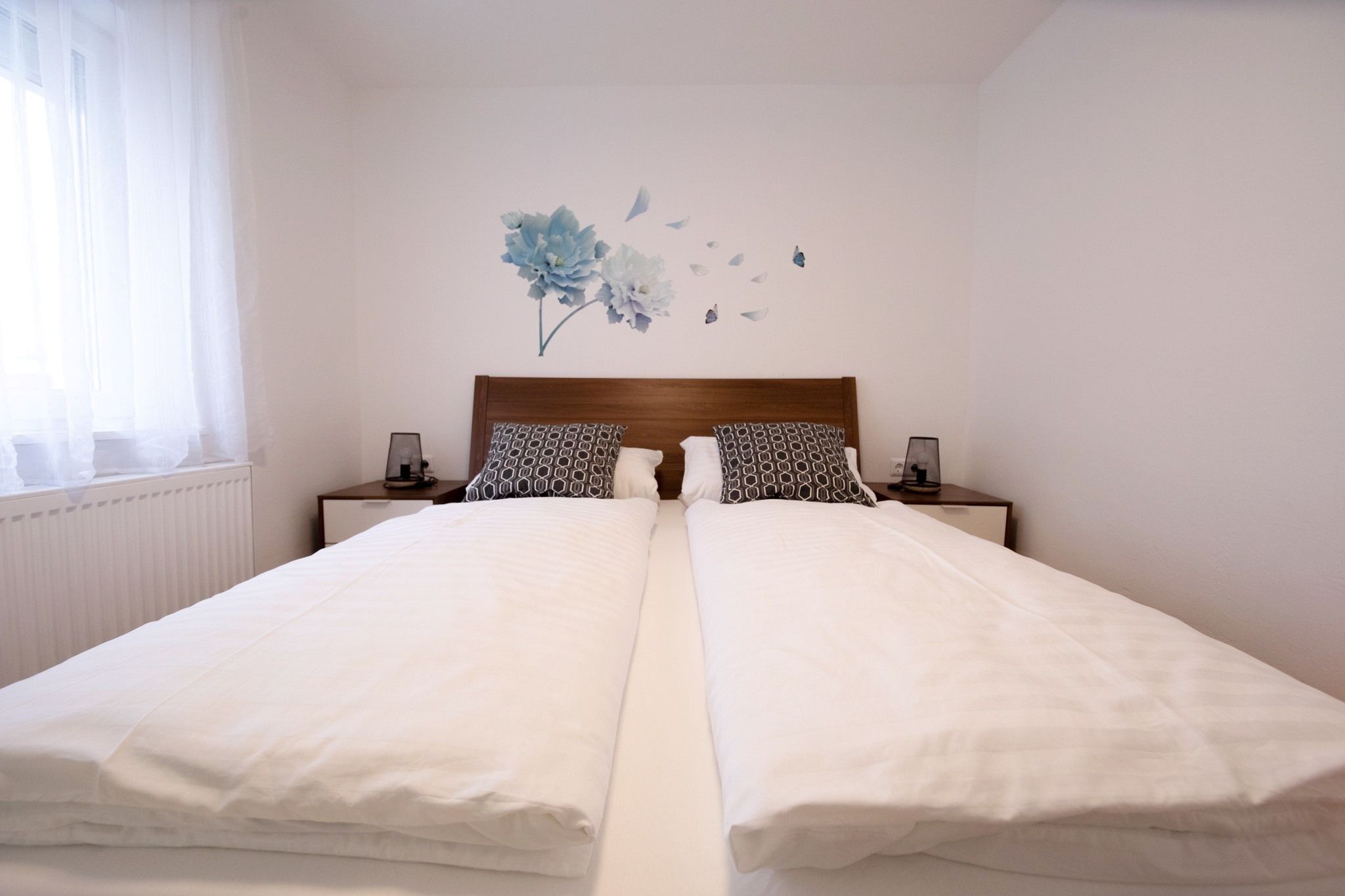 A modern bedroom with a double bed, white comforters and decorative pillows. There is a floral motif on the wall.