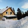 A yellow house with a snow-covered roof and balcony, surrounded by trees and snow.