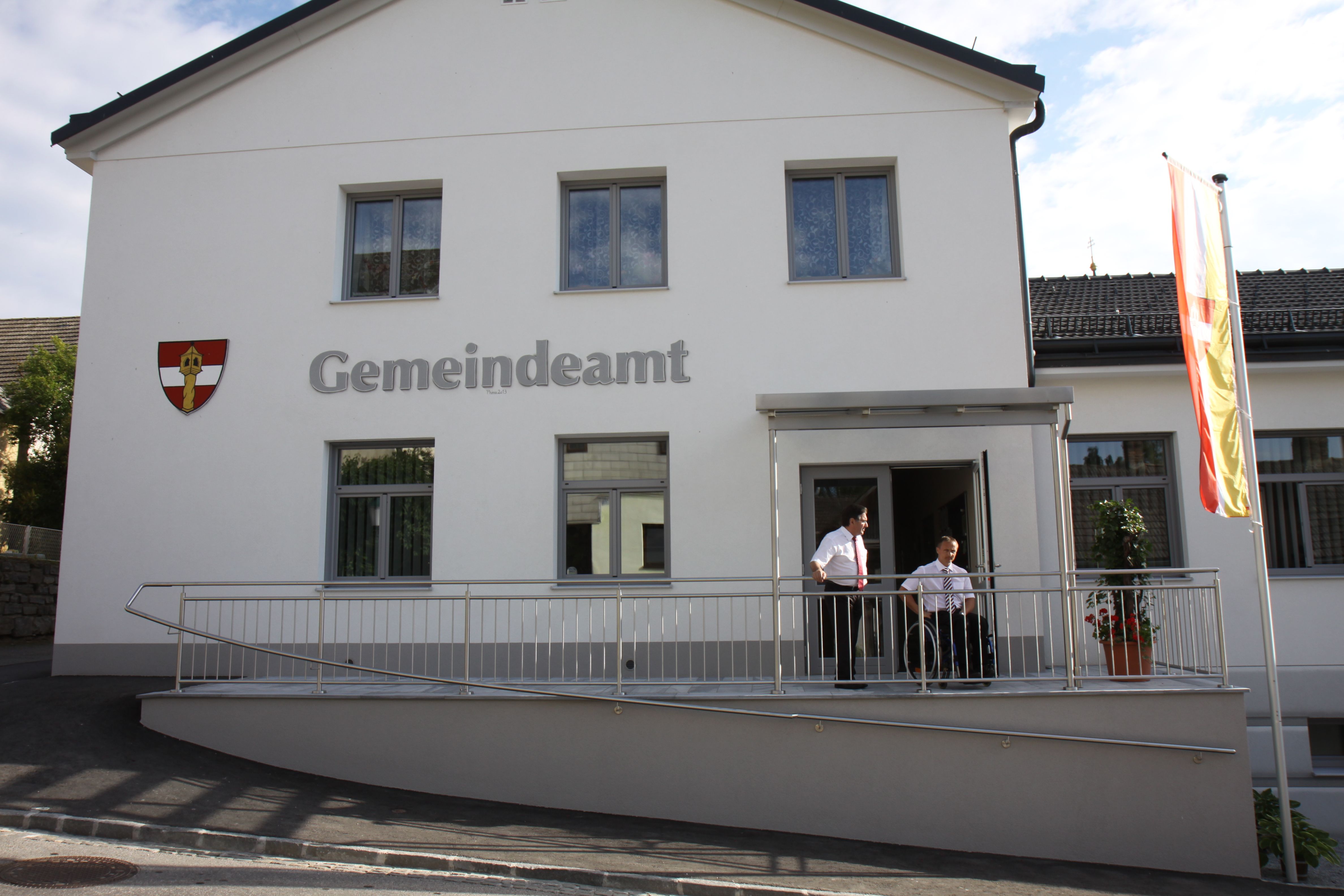 A modern municipal office with a white façade, two people standing on a ramp, a flag flying next to it.