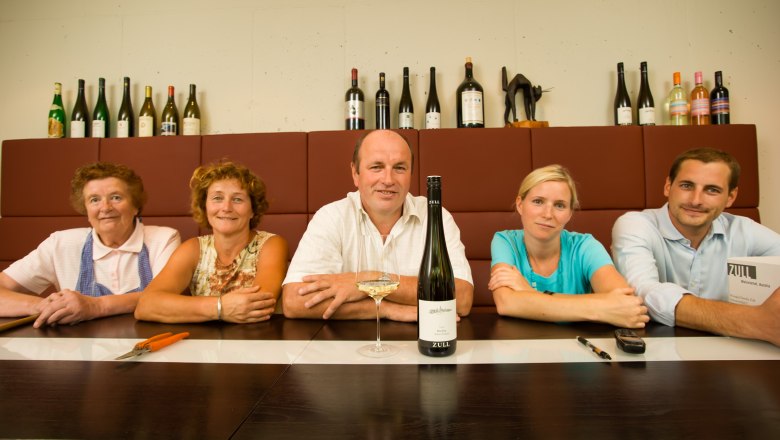 Five people sit at a table with bottles of wine in the background.