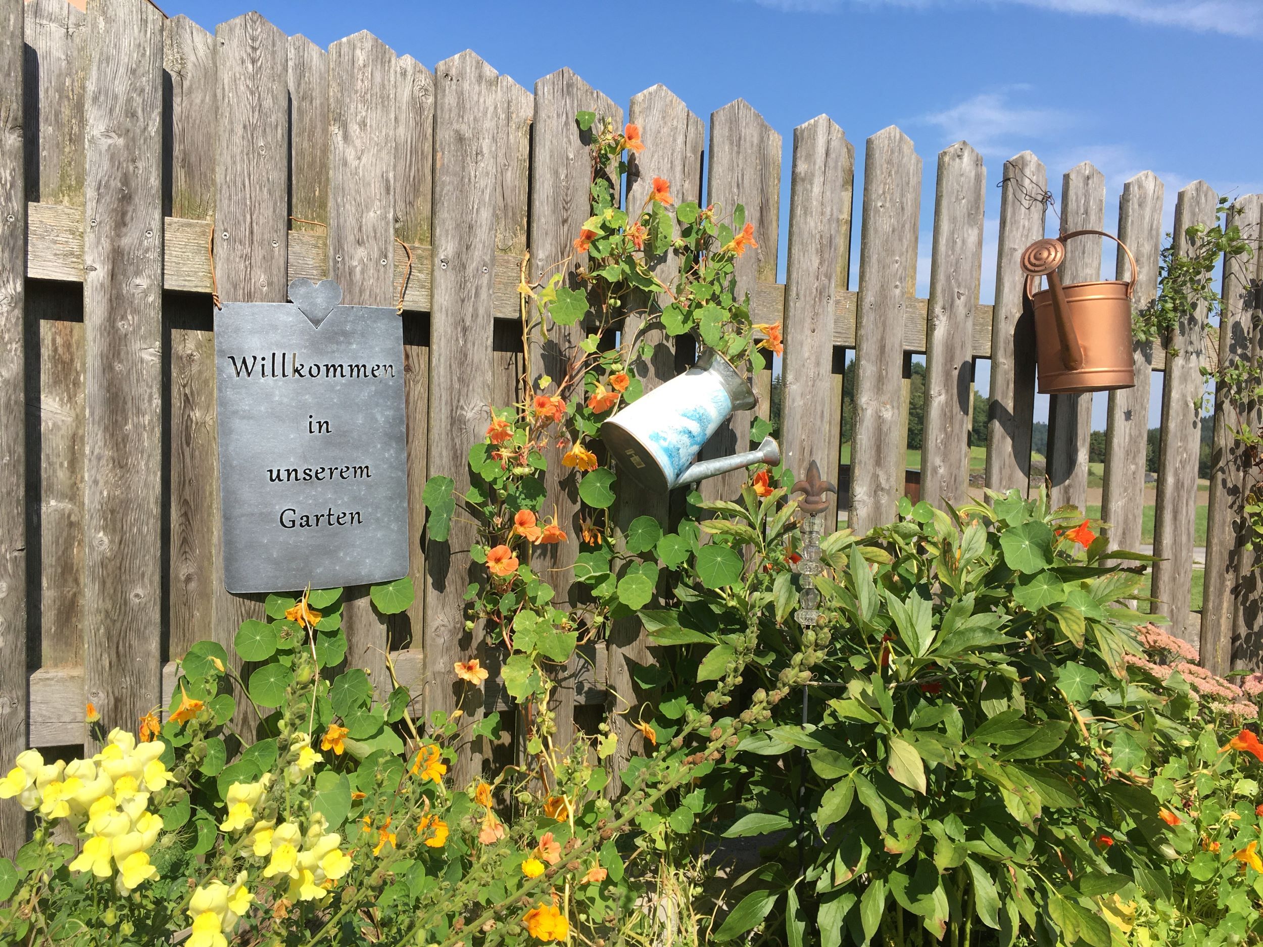 A wooden fence with a 'Welcome to our garden' sign, surrounded by flowers and watering cans.
