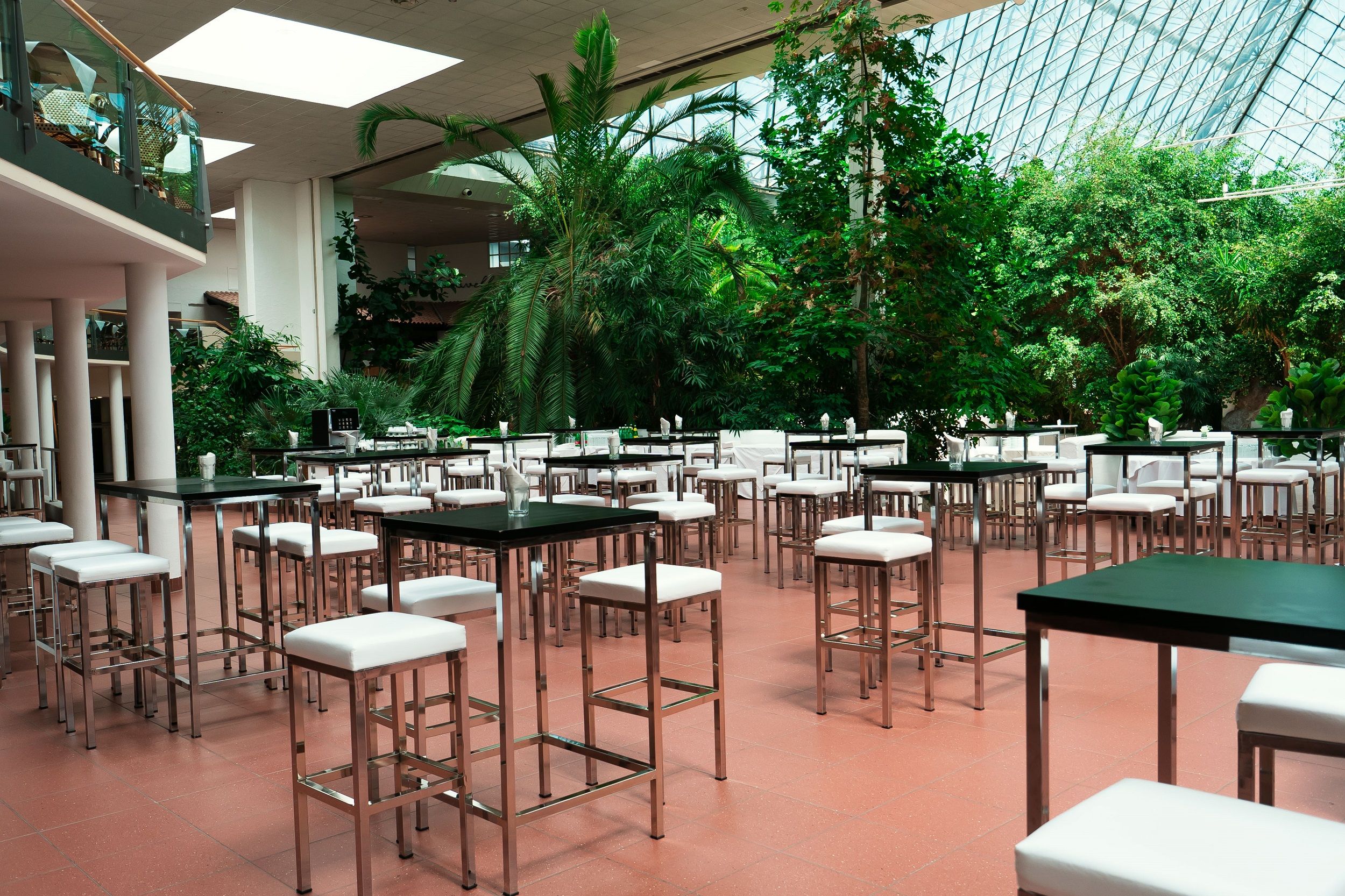 Interior view of a modern event room with high tables and chairs, surrounded by lush plants and a glass roof.