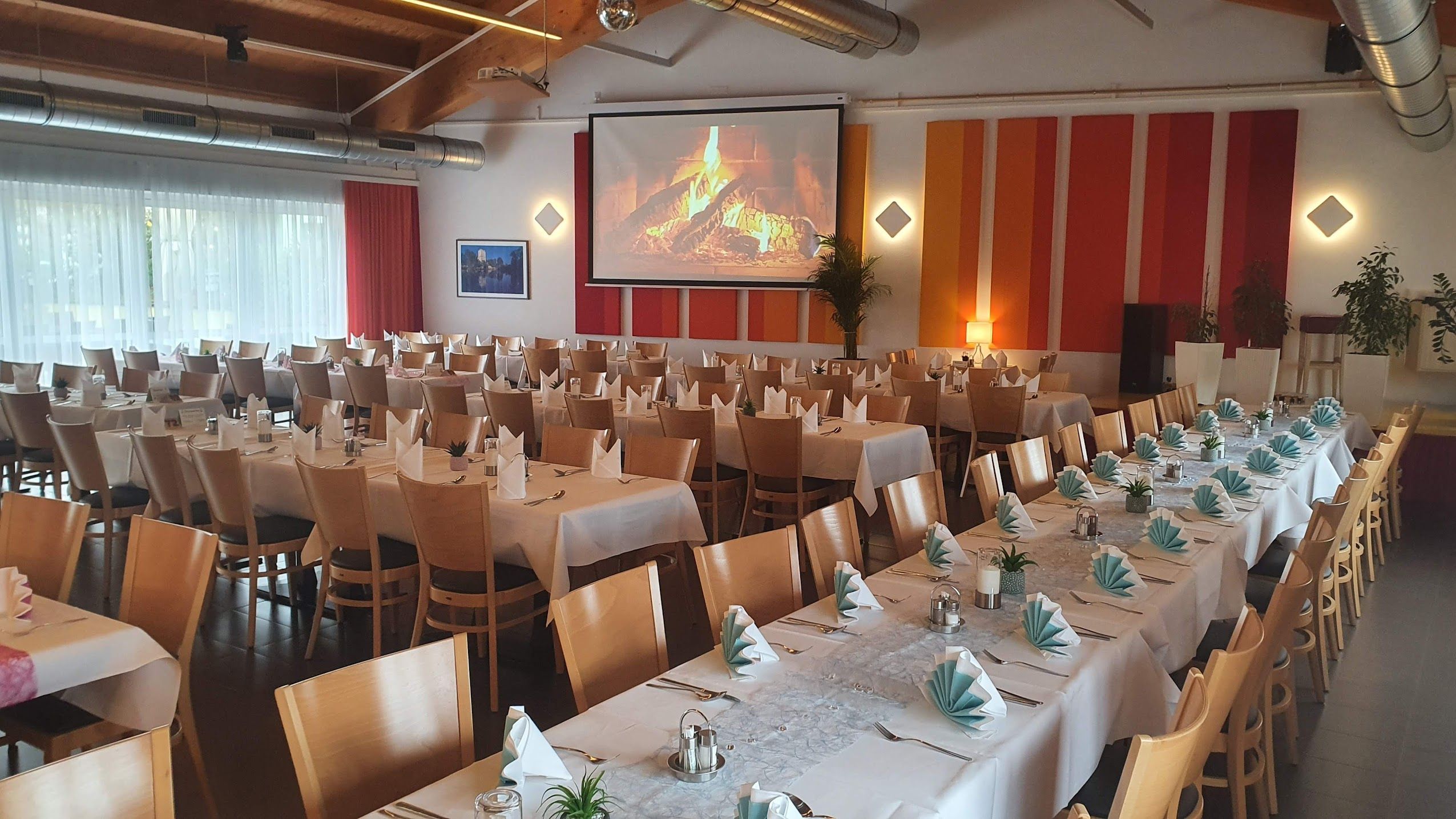 A festively decorated dining room with long tables, white tablecloths and folded napkins. On the wall, a screen with a log fire.