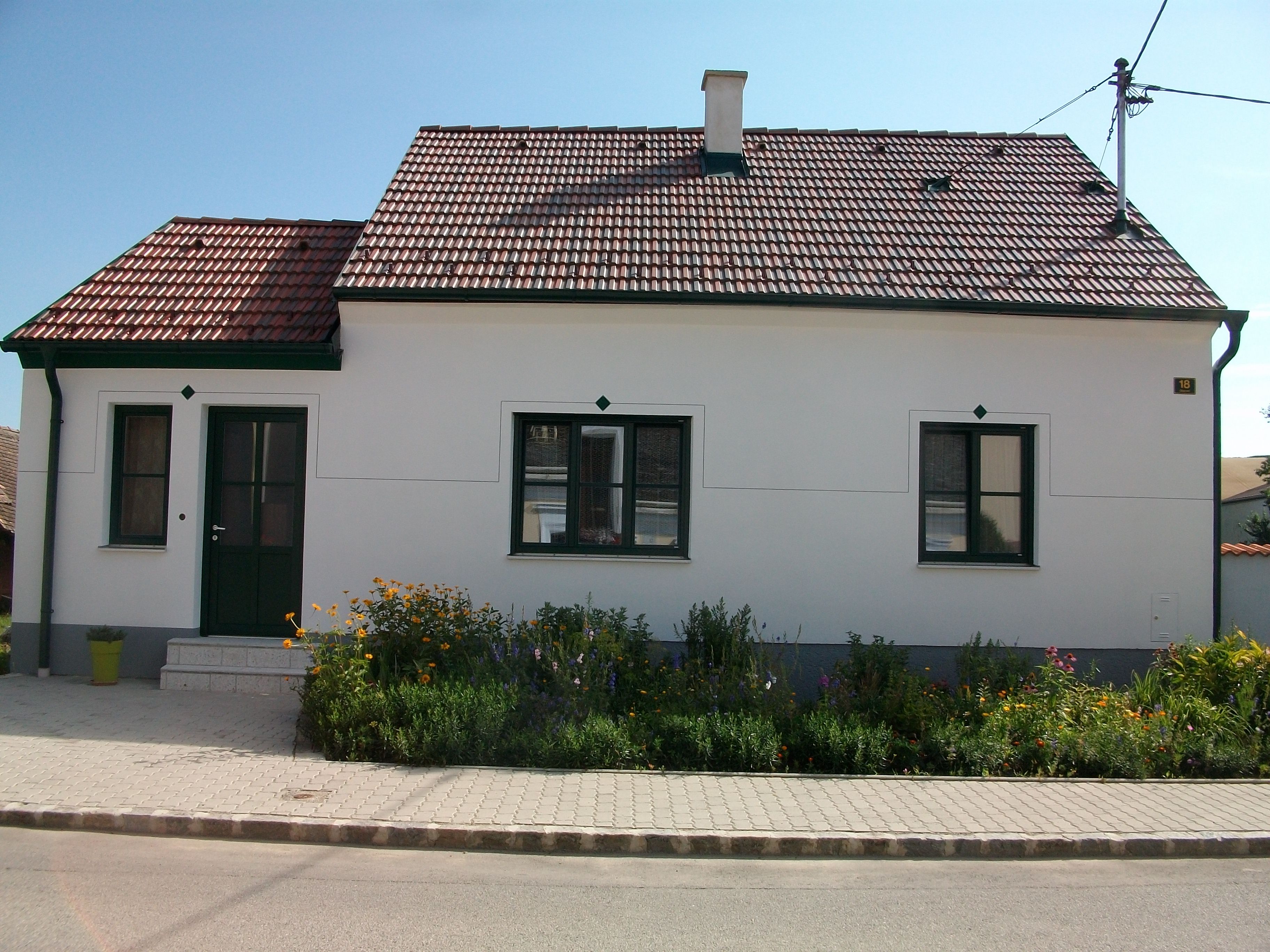 A small, white house with a red tiled roof and green door frame, surrounded by a well-tended garden with flowers.