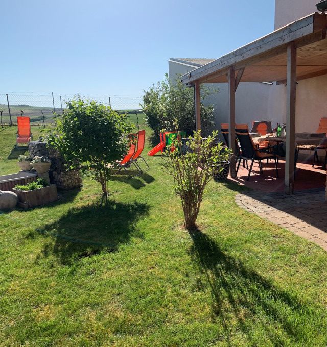 A sunny garden with lawn, shrubs, sun loungers and a covered terrace with table and chairs.