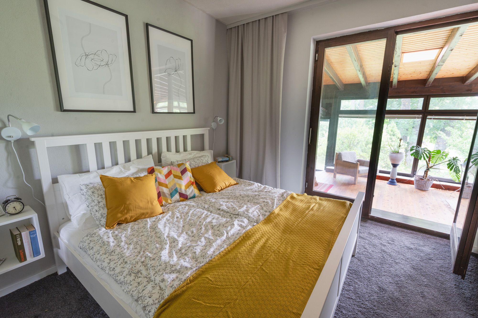 A cozy bedroom with a white bed, yellow cushions and a terrace with plants in the background.
