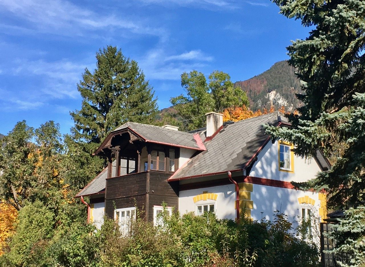 A picturesque country house with a gray roof and yellow window frames, surrounded by trees and mountains in the background.
