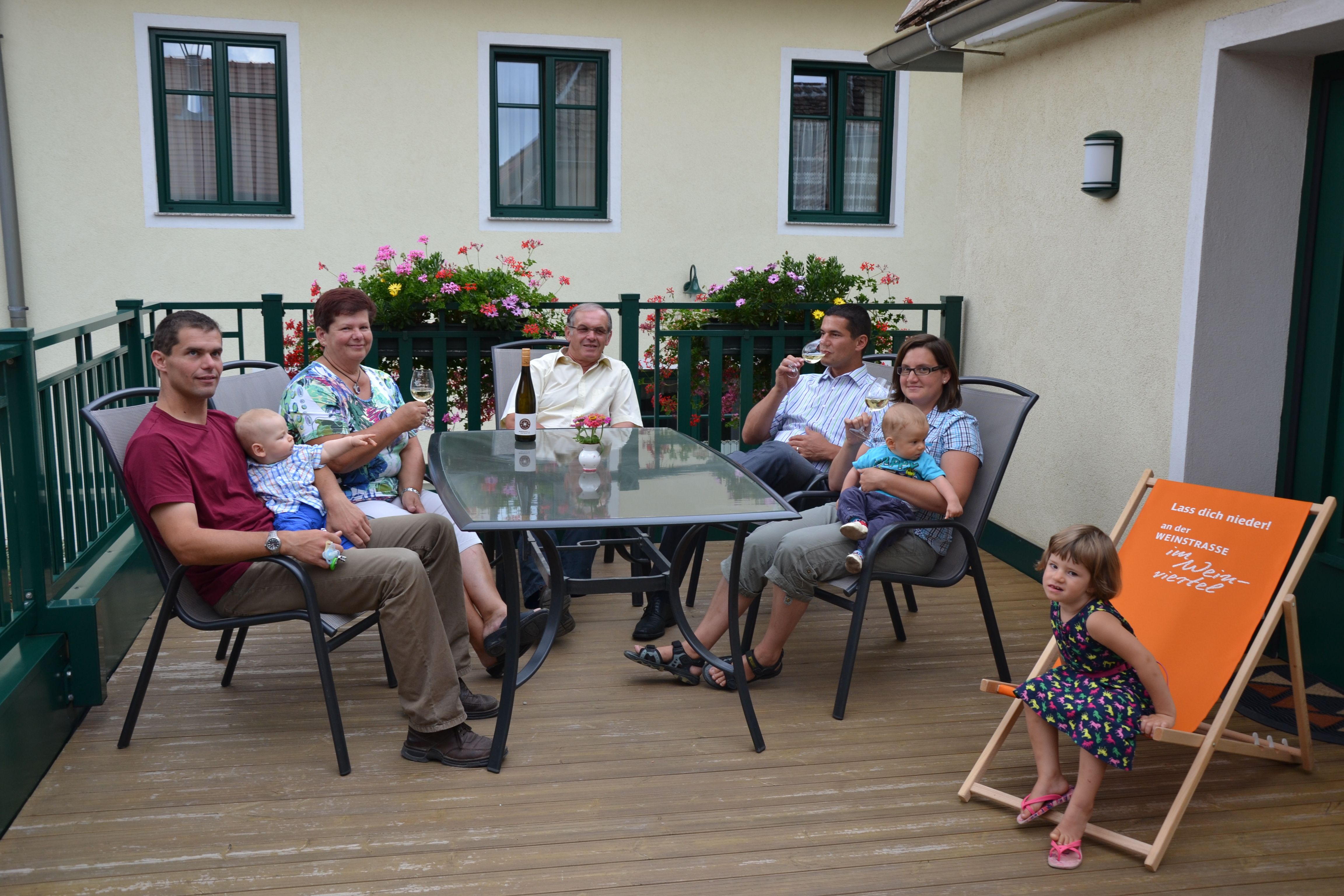 A family is sitting on a terrace at a table with drinks. Flowers and a building can be seen in the background.