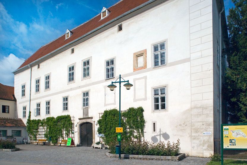 Traismauer Castle with a white façade and red roof tiles, surrounded by plants and a paved forecourt.