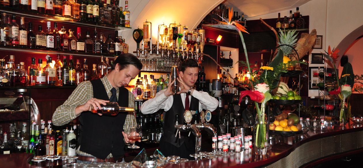 Two bartenders work in a stylish bar with lots of bottles in the background.