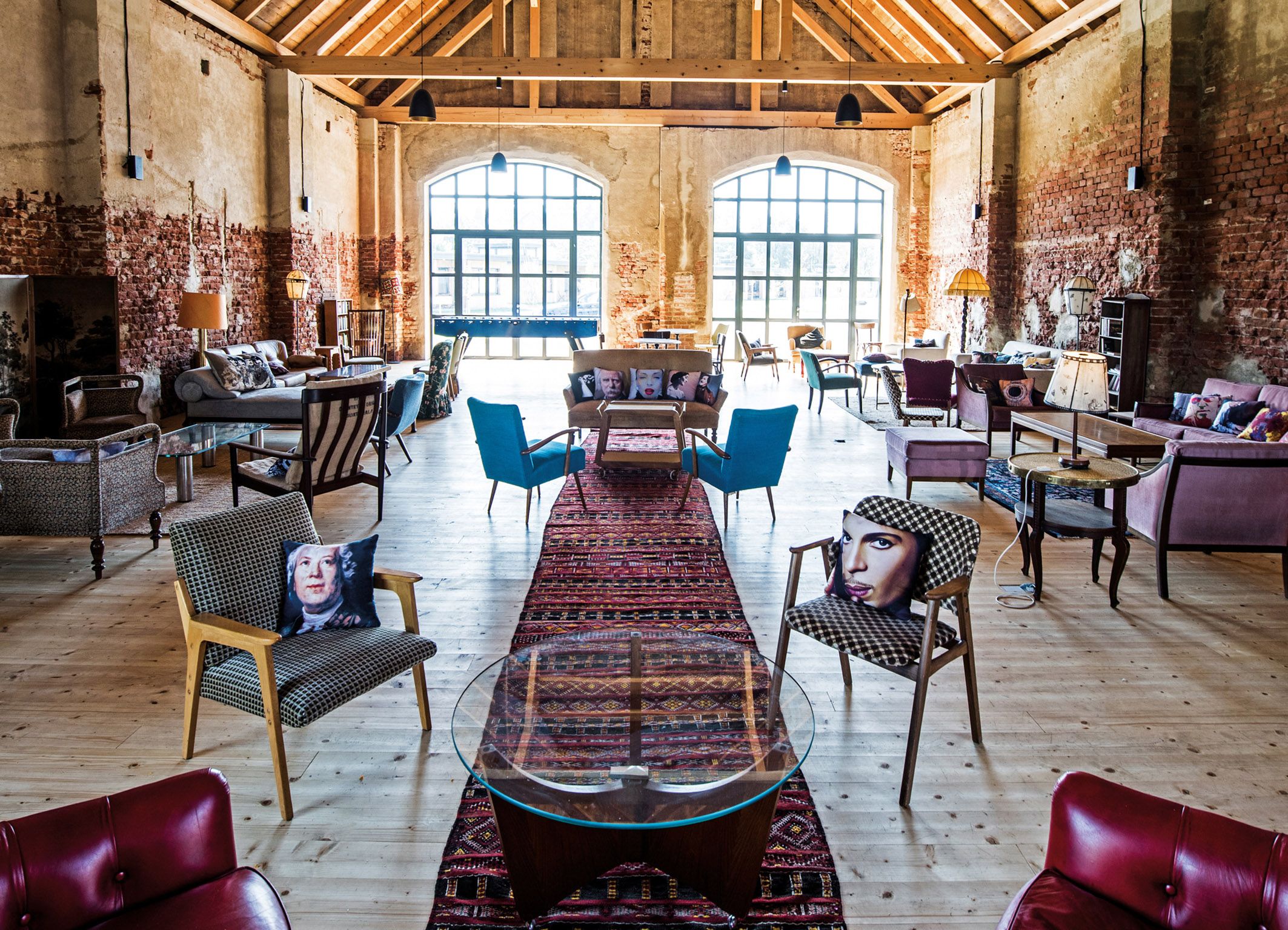 Interior view of a stylishly furnished barn with wooden floor, brick walls and large windows. Various pieces of seating furniture and colorful cushions can be seen.