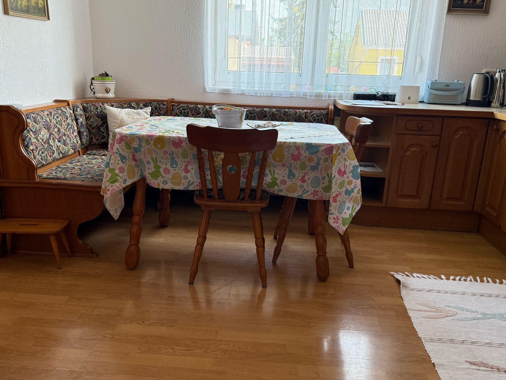 Dining area with wooden table, chairs and corner bench, colorful tablecloth, window in the background.