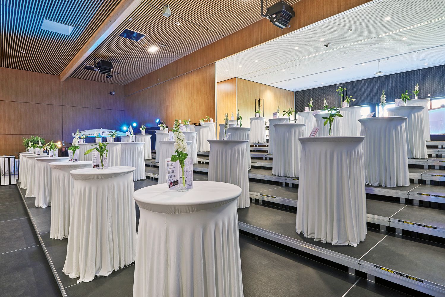 An event room with high bar tables covered in white, decorated with flowers and information material.
