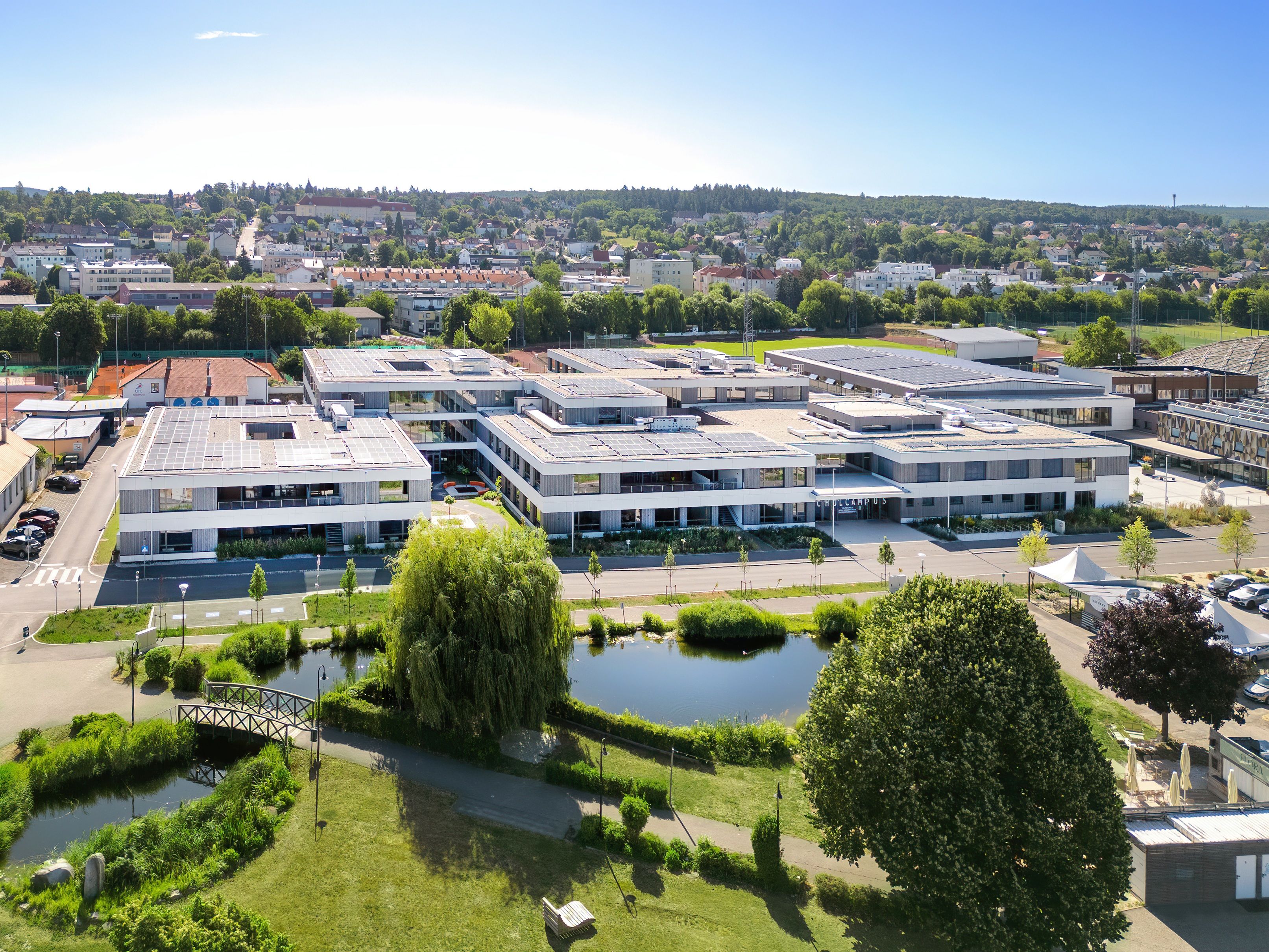 Aerial view of a modern school campus with solar panels on the roofs, surrounded by green spaces and a pond in the foreground.