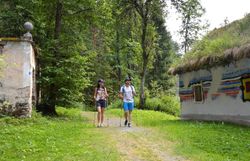 Ein Paar wandert entspannt auf einem schmalen Pfad, umgeben von üppigem Grün und der beruhigenden Stille des Waldes. Die bunten, kunstvoll gestalteten Wände der Hundertwassermühle verleihen der Umgebung einen einzigartigen Charme und laden dazu ein, die kreative Verbindung von Natur und Kunst zu entdecken.