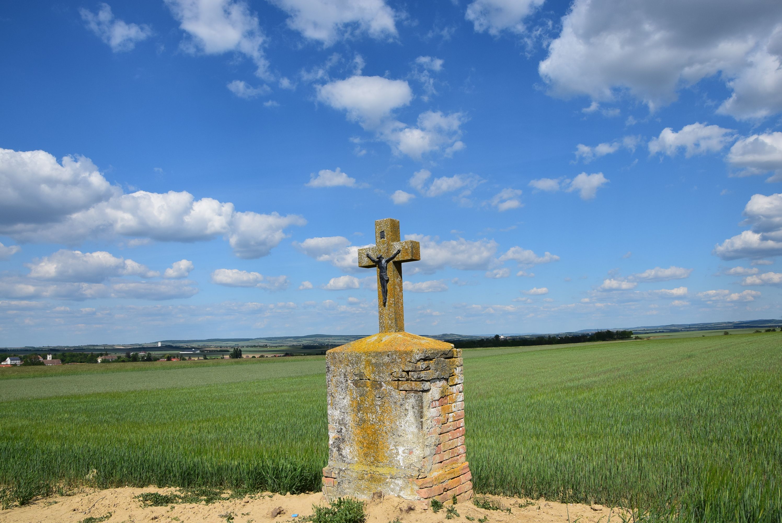 A stone cross with a figure of Christ stands in a field under a blue sky with clouds.