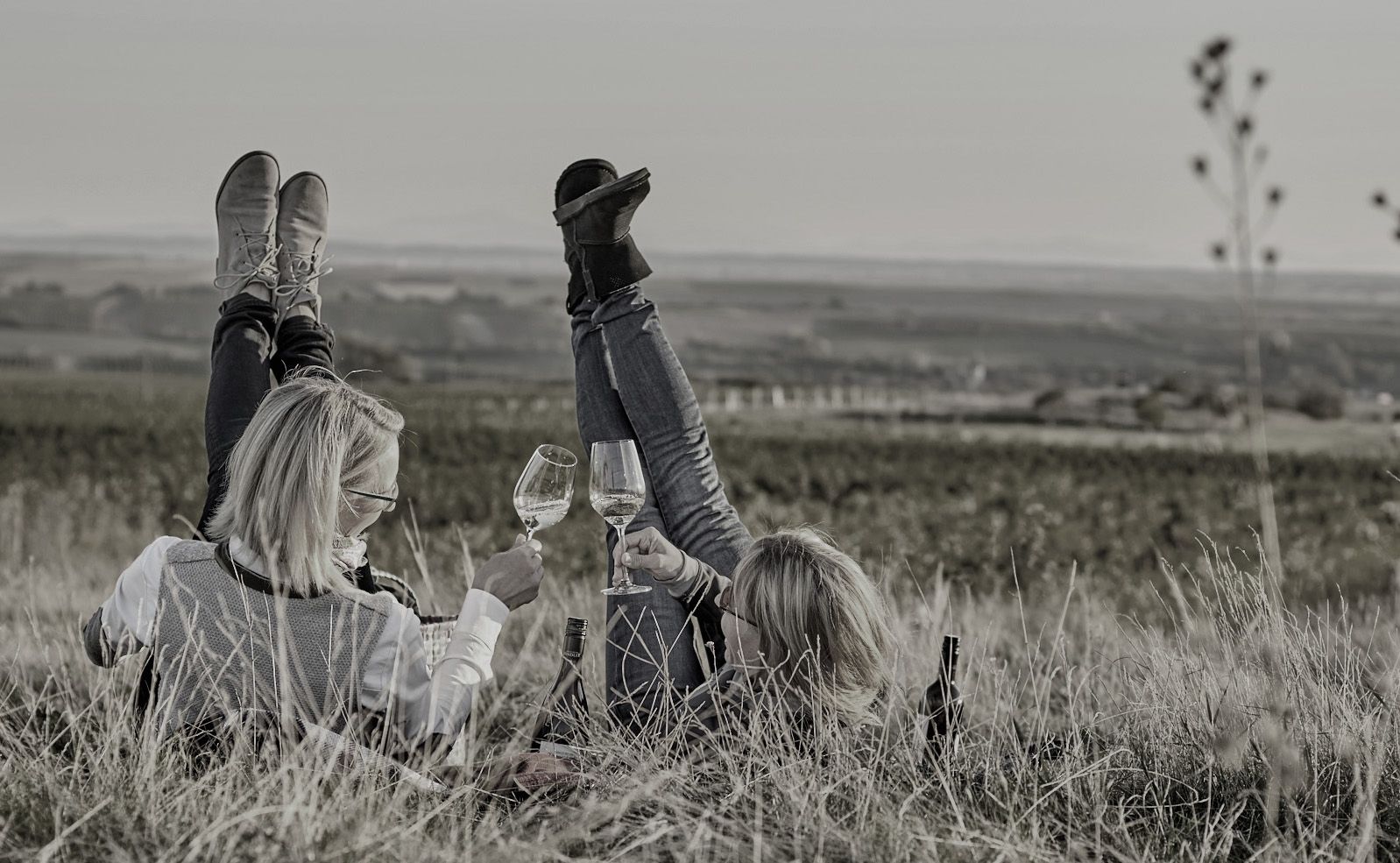 Two people are lying on the grass with wine glasses in their hands, legs stretched up in the air, in front of a vast landscape.