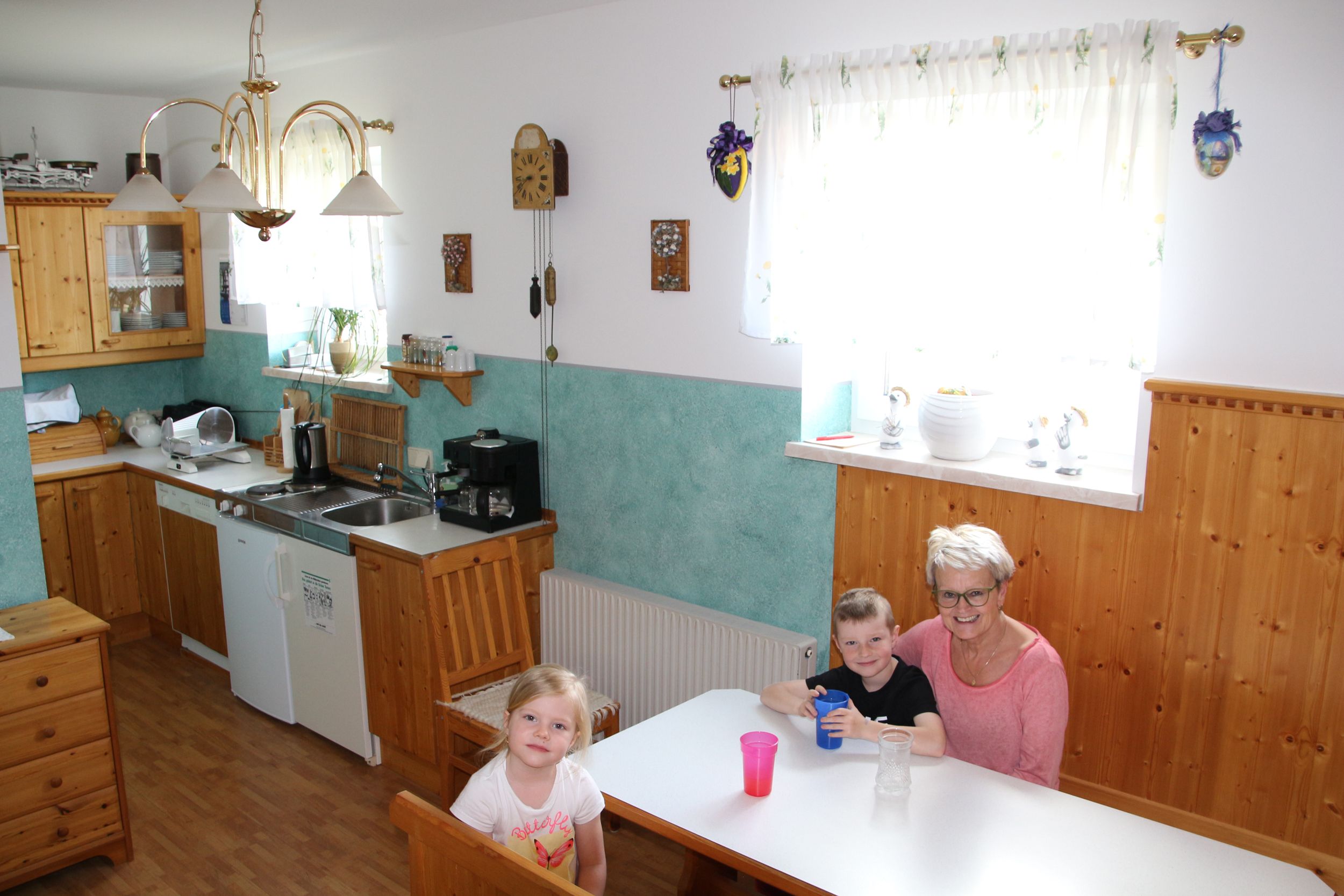 A breakfast room with wooden furniture, a kitchen in the background and three people at a table.