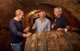 Three men in a wine cellar tasting wine.