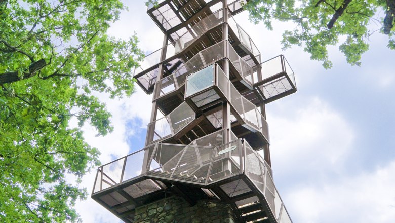 A modern observation tower made of metal and stone, surrounded by green trees, rises into the blue sky.