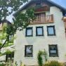 A traditional house with white walls, wooden cladding and a balcony, surrounded by green trees and plants.