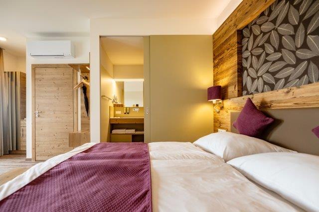 Modern furnished hotel room with double bed, wooden walls and view into the bathroom.