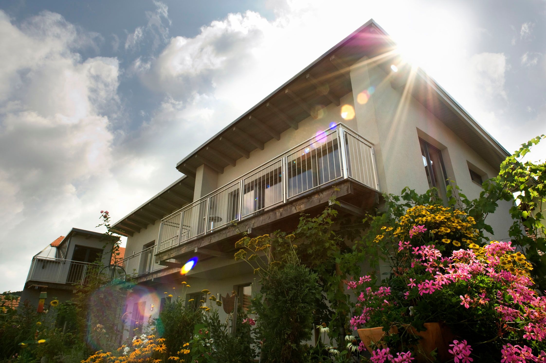 Exterior view of a house with balcony and flowering garden in the foreground, rays of sunshine in the sky.