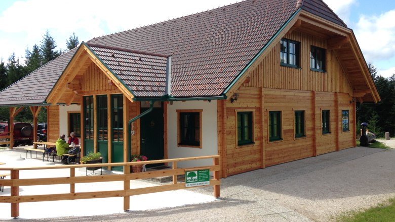 A wooden hut with a green roof and windows, surrounded by trees. Two people are sitting at a table outside.