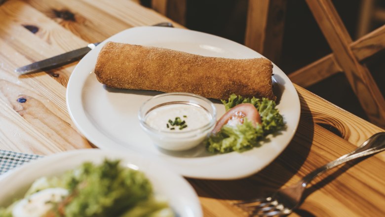 A plate with a breaded, baked dish, salad garnish and a bowl of dip on a wooden table.