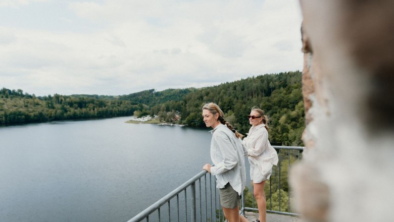 Two women stand on a balcony with a view of a lake and wooded hills.