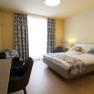 Modernly furnished hotel room with double bed, desk, TV and armchairs.