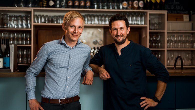 Two men stand smiling at a bar, surrounded by glasses and bottles.