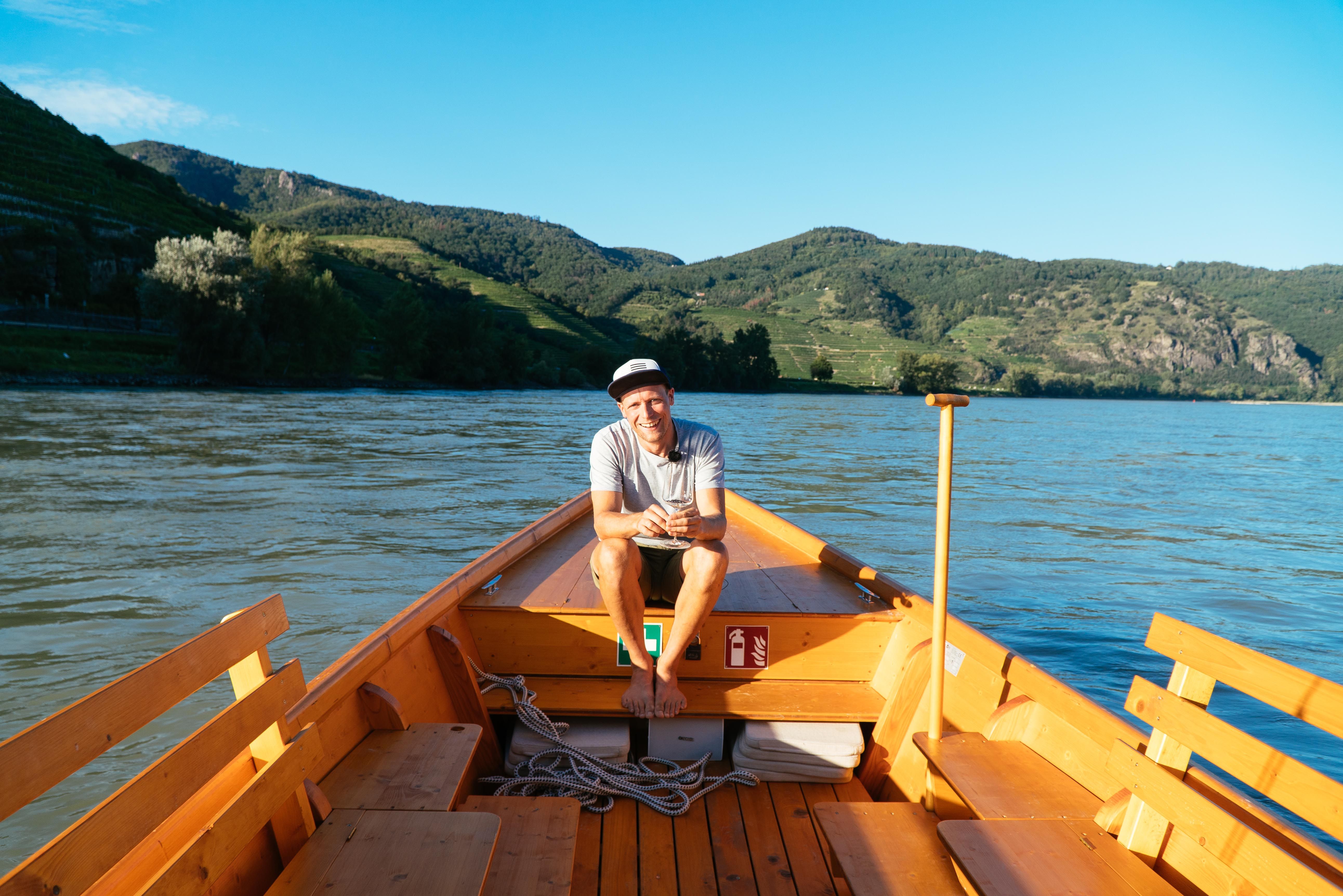 Person sitting on a wooden boat on a river, surrounded by green hills.