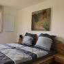 A cozy bedroom with a double bed, grey bed linen, a painting on the wall and a window with blinds.