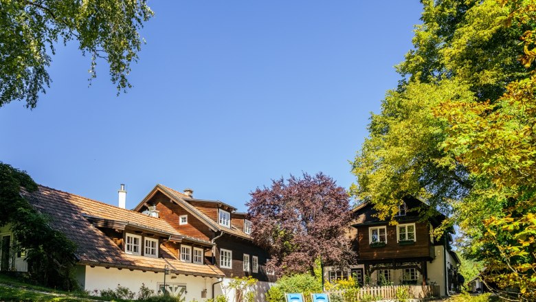 A traditional country house with a wooden roof and blue deckchairs in the garden, surrounded by trees and blue skies.