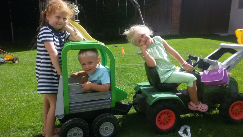 Three children playing in the garden with a toy tractor and trailer.