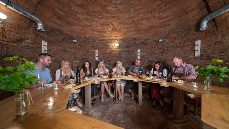 Group of people sitting at a wooden table in a wine cellar.