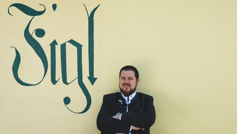 A man in traditional dress stands in front of a yellow wall with the word 'Figl' written on it.