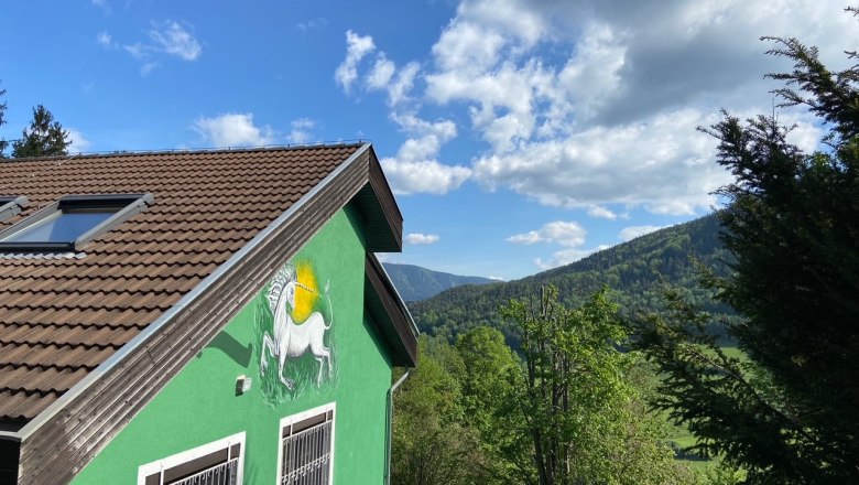 Unicorn house, © Wieneralpen Green house with unicorn picture on the wall, surrounded by trees and mountains under a blue sky.