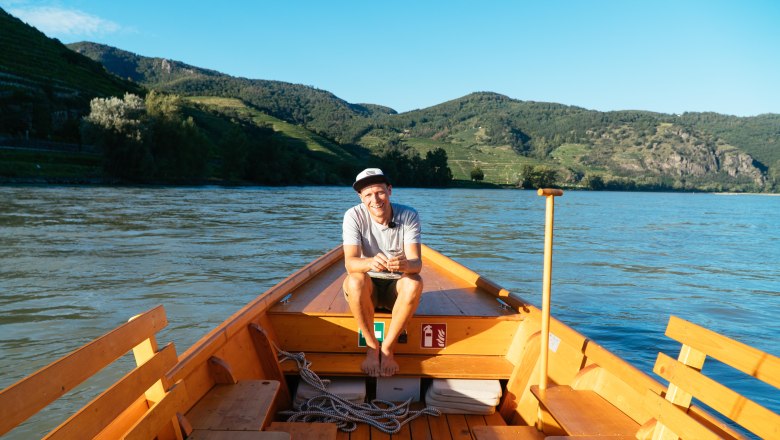Person sitting on a wooden boat on a river, surrounded by green hills.