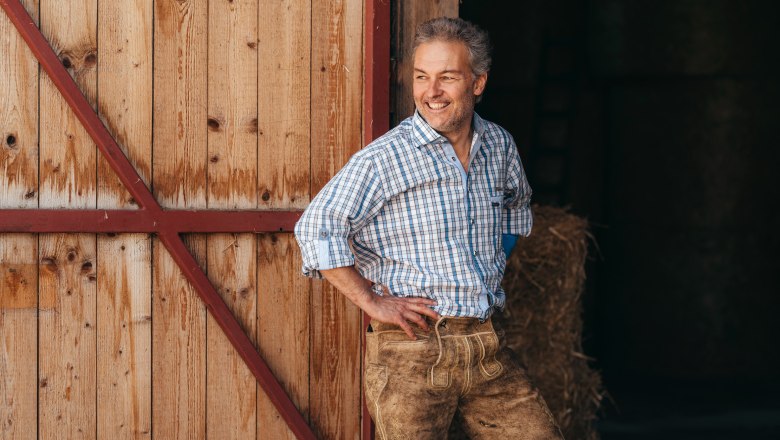 Man in plaid shirt and leather pants leans against wooden wall, smiles.