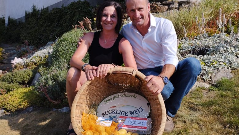 A woman and a man sit smiling in a garden, holding a basket of potatoes and eggs.