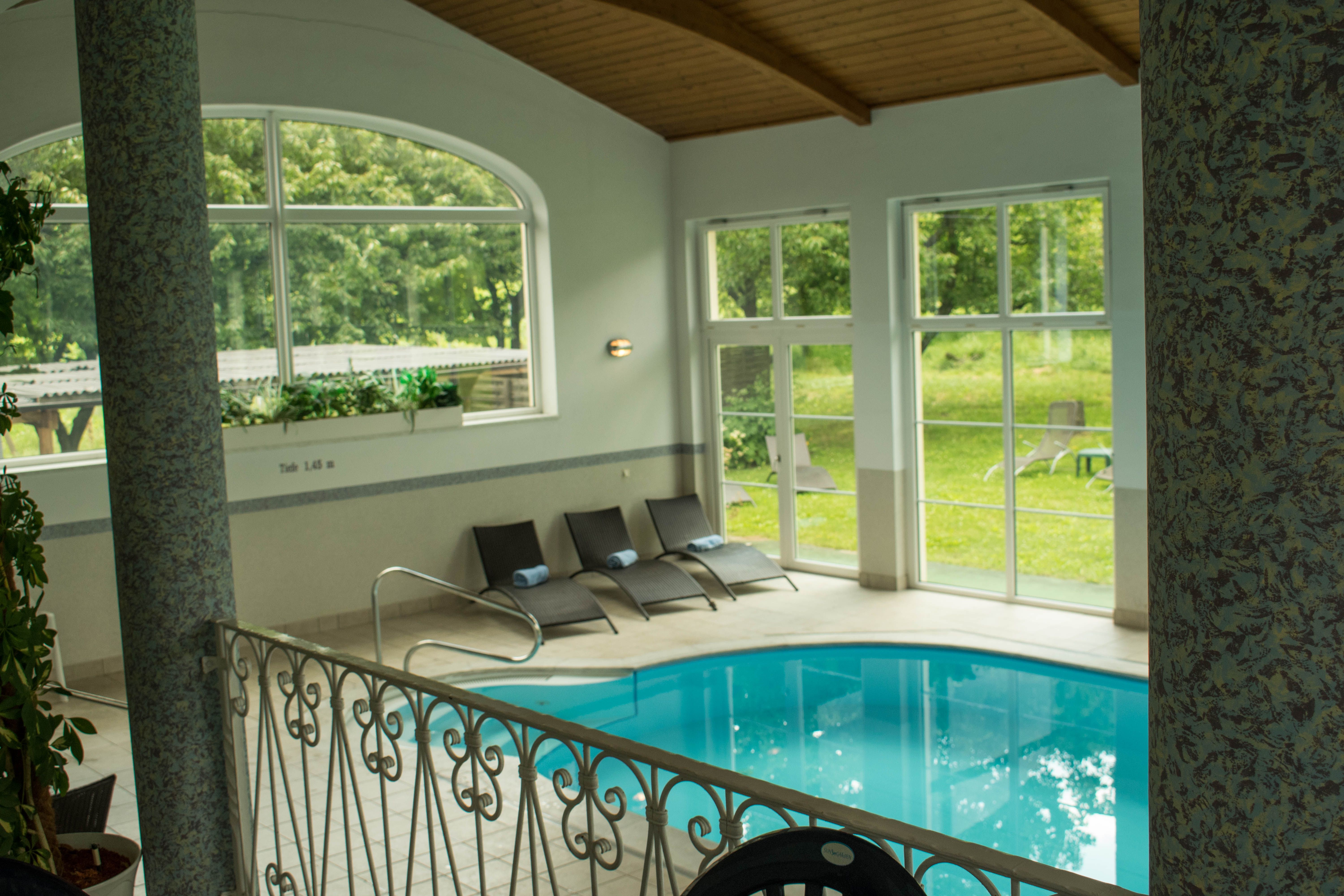 Interior view of a hotel pool with sun loungers and large windows overlooking the greenery.