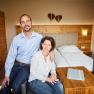 A couple is sitting in a cozy room with wooden furniture and a double bed.