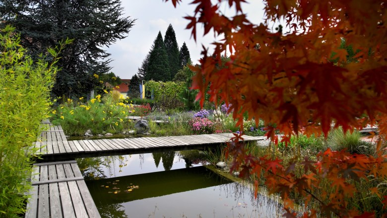 A garden with wooden walkways over a pond, surrounded by colorful flowers and trees in autumn leaves.