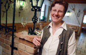A smiling person holds a glass of white wine in a cozy room with a brick wall and a lantern.