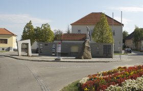 Monument in Mailberg with statue on a rock, surrounded by buildings and flowerbeds.